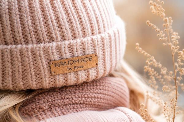 Prostokątna jasnobrązowa metka ze sztucznej skóry "Handmade by Bine" na różowej czapce.